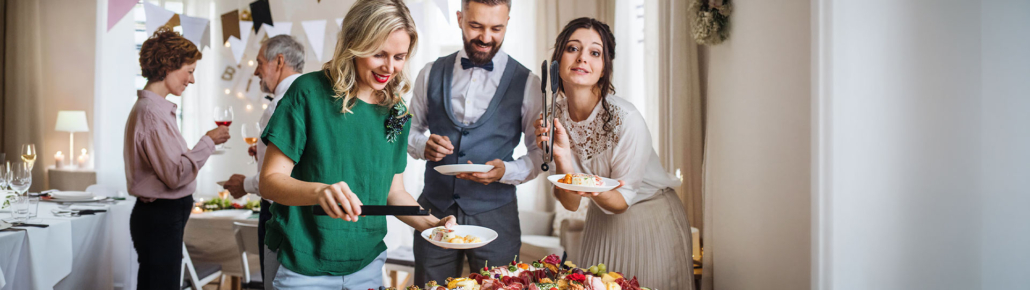 Drei elegant gekleidete Gäste vor einem Buffet stehend und lächelnd. Sie wählen ihre Fingerfood Snacks aus und sind gut drauf.