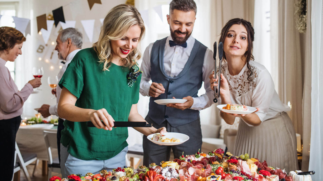 Drei elegant gekleidete Gäste vor einem Buffet stehend und lächelnd. Sie wählen ihre Fingerfood Snacks aus und sind gut drauf.