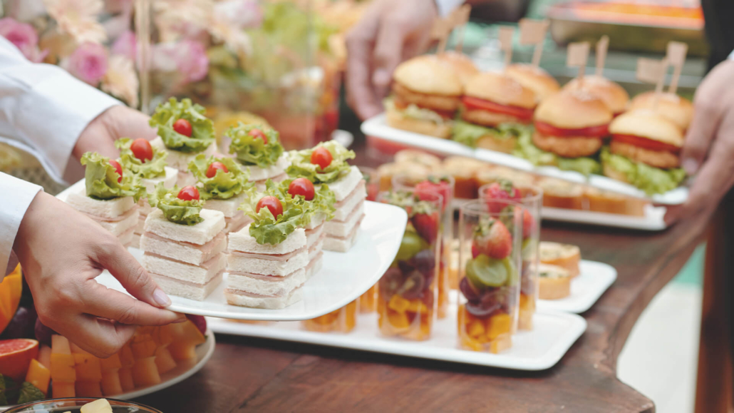 Bildauschnitt eines Buffettisches. Im Vordergrund sieht man einen Teller, der von Frauenhänden gehalten wird auf dem gestapelte Sandwichtoast-Häppchen zu sehen sind, sowie ein Teller gehalten von Männerhänden, auf dem Burger mit Hähnchenschnitzel zu sehen sind, die mit einem Fähnchenspieß dekotiert sind. Die Teller werden gerade auf dem Tisch abgestellt.