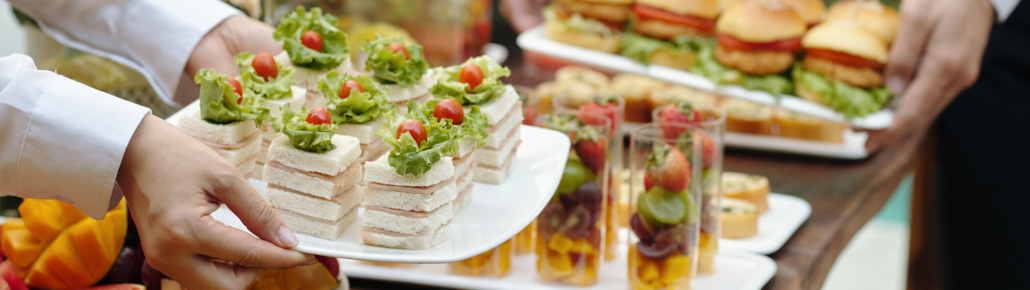 Bildauschnitt eines Buffettisches. Im Vordergrund sieht man einen Teller, der von Frauenhänden gehalten wird auf dem gestapelte Sandwichtoast-Häppchen zu sehen sind, sowie ein Teller gehalten von Männerhänden, auf dem Burger mit Hähnchenschnitzel zu sehen sind, die mit einem Fähnchenspieß dekotiert sind. Die Teller werden gerade auf dem Tisch abgestellt.